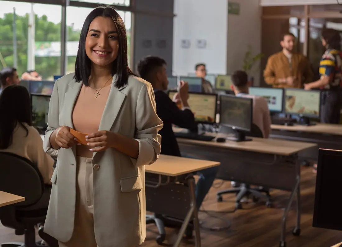 Profesional de tecnología sonriendo en una oficina con su equipo trabajando al fondo
